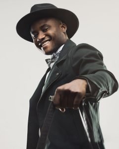 Portrait of handsome young man in black suit and hat holding handbag. Happy young male traveler looking at camera and smiling. Vertical studio photography from a DSLR camera. Sharp focus on eyes.